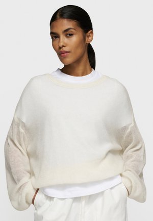 Lichtgewicht gebreide trui met een relaxte pasvorm, doorschijnende mouwen en een ronde halslijn, gedragen over een witte t-shirt. Gevoel van textuur.