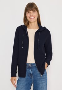 Donna sorridente che indossa una felpa con zip blu navy sopra una maglietta beige e jeans blu, in piedi contro uno sfondo chiaro e uniforme.