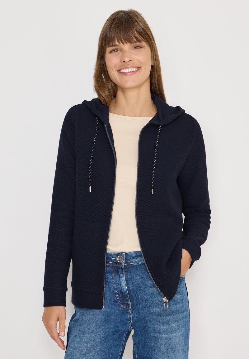 Donna sorridente che indossa una felpa con zip blu navy sopra una maglietta beige e jeans blu, in piedi contro uno sfondo chiaro e uniforme.