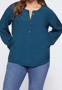 Teal long-sleeve blouse with a rounded neckline, button placket, and a relaxed fit. Fabric has a smooth texture, paired with light blue jeans.