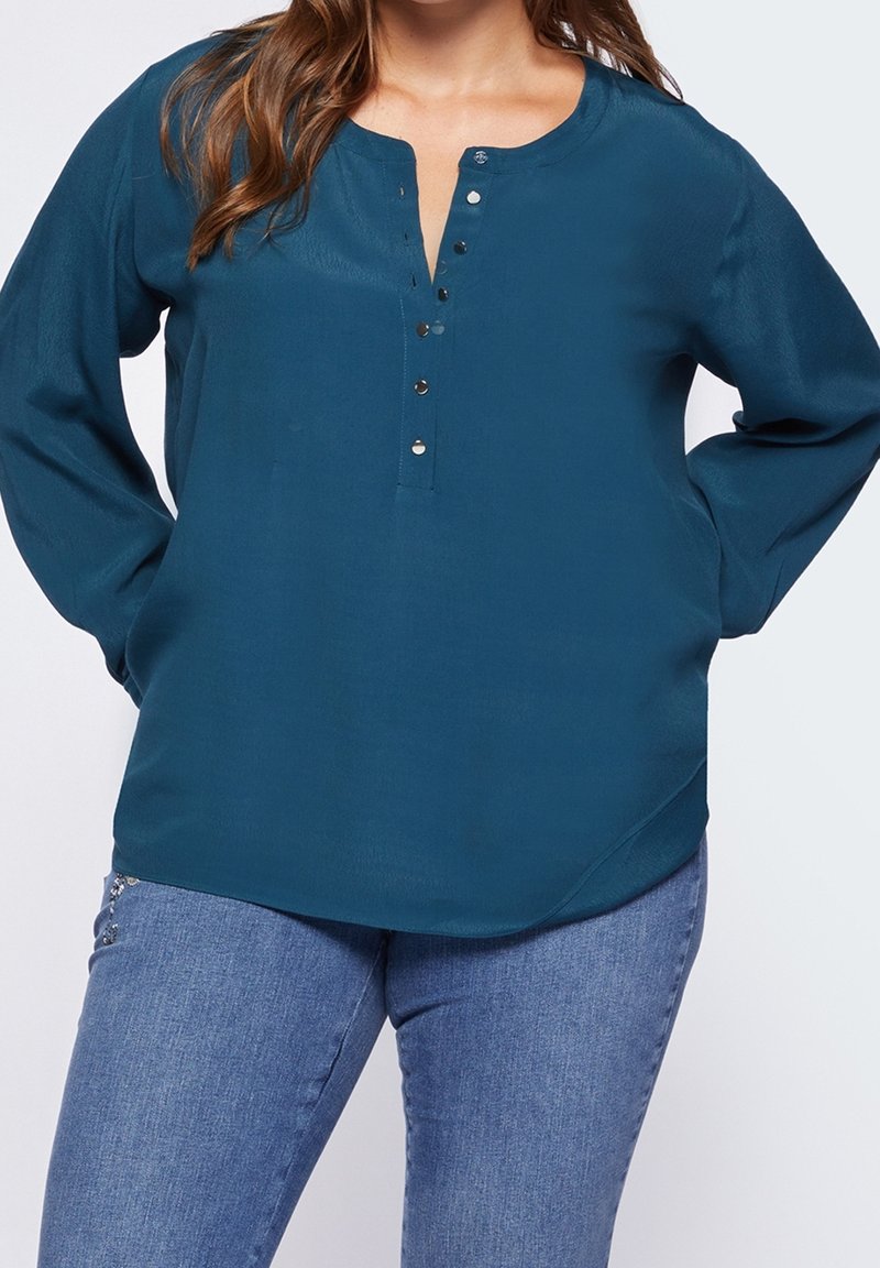 Teal long-sleeve blouse with a rounded neckline, button placket, and a relaxed fit. Fabric has a smooth texture, paired with light blue jeans.