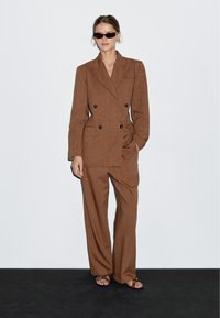 Brązowy lniany marynarka podwójnie zapinana z dużymi guzikami, w zestawie z szerokimi spodniami o pasującym kolorze. Modelka nosi małe czarne okulary przeciwsłoneczne i sandały na cienkich paskach.