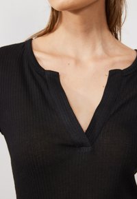 Haut noir à nervures avec un décolleté en V et des manches courtes, en tissu texturé qui semble légèrement transparent. Gros plan sur le décolleté.