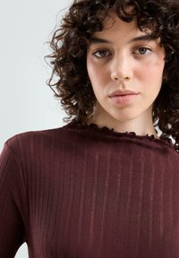 Bordeaux geribbelde top met hoge hals en een ruchesrand. Zachte textuur en aansluitend ontwerp, met verticale strepen.