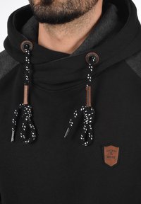 Zwarte hoodie met grijze accenten, voorzien van een hoge kraag, leren label en een getextureerd stoffen ontwerp. De trekkoorden hebben een patroon en metalen tips.