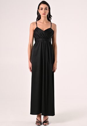 Schwarzes, bodenlanges Kleid aus glattem Stoff, das ein tailliertes Oberteil mit dünnen Trägern, eine geraffte Taille und einen fließenden Rock hat. Schwarze Riemchen-Highheels.