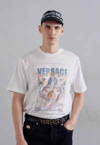 Balta trumpa rankovė T-palaidinė su spalvinga Versace grafika, vaizduojančia jūros motyvus. Suderinta su tamsiomis džinsinėmis kelnėmis ir juodu diržu su auksine sagtimi.