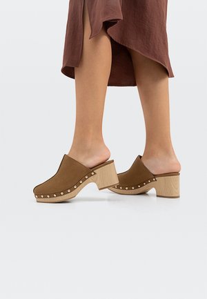 Bruine suède klompen met een hoge, gestructureerde houten hak en decoratieve metalen studs langs de zijkant. Open achterkant ontwerp, minimalistische vorm.