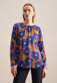 Blusa a maniche lunghe color senape con motivo floreale blu, dotata di dettagli bianchi e scollatura arricciata. Tessuto morbido con una vestibilità comoda.