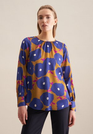 Blusa a maniche lunghe color senape con motivo floreale blu, dotata di dettagli bianchi e scollatura arricciata. Tessuto morbido con una vestibilità comoda.