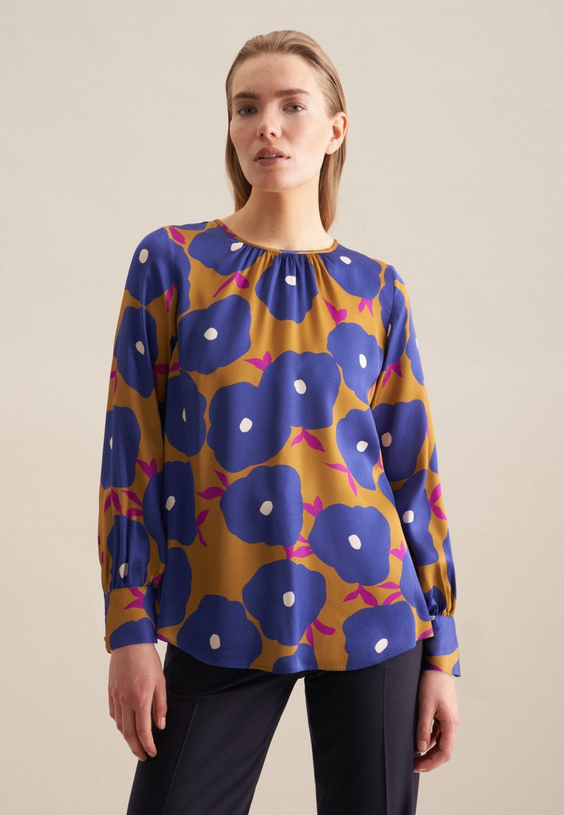 Blusa a maniche lunghe color senape con motivo floreale blu, dotata di dettagli bianchi e scollatura arricciata. Tessuto morbido con una vestibilità comoda.