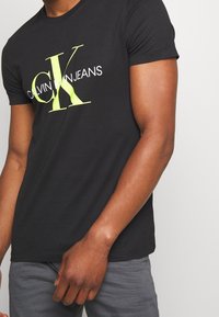 Mężczyzna w czarnej koszulce Calvin Klein Jeans z dużymi żółtymi literami "CK" i białym tekstem, w połączeniu z szarymi spodniami.