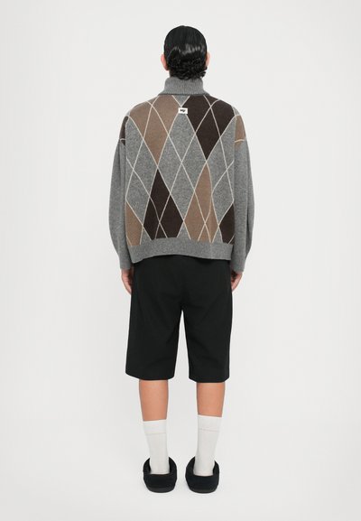 Suéter gris de punto con patrón de diamante en tonos marrones, con cuello alto. Combinado con pantalones cortos negros, calcetines blancos y zapatillas negras.