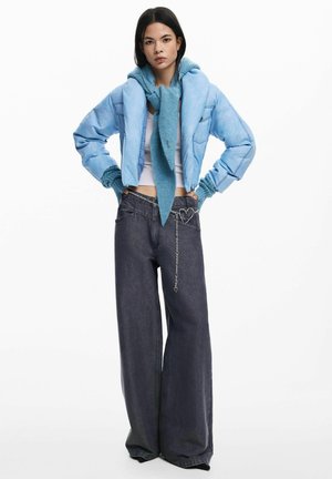 Vrouw draagt een lichtblauwe gewatteerde jas met bijpassende sjaal, wit cropped topje, wijde donkere jeans en een zilveren kettingriem met hartvorm.