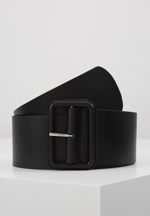 Large ceinture en cuir noir avec une grande boucle rectangulaire revêtue reposant sur une surface blanche sur un fond neutre.