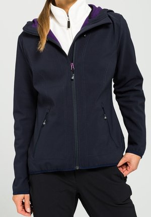 Kobieta w granatowej kurtce na zamek z fioletową podszewką kaptura i białym pod spodem, trzymająca brzeg kurtki jedną ręką.