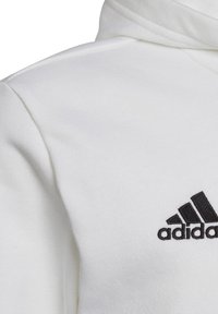 Hvid sweatshirt med et struktureret stof, som har et sort broderet Adidas-logo på brystet. Indeholder en hætte og syede sømme.