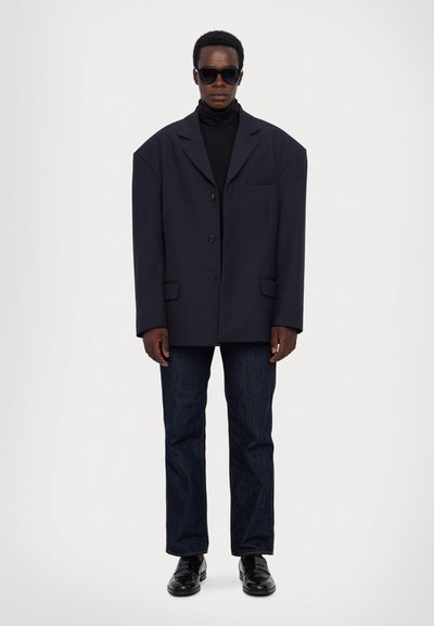 Marineblauer oversized Blazer, einreihig mit zwei Knöpfen, seitlichen Taschen, getragen über einem schwarzen Rollkragenpullover. Dunkelblaue Jeans und schwarze Schuhe.