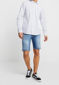 Muž v bílo-modrém pruhovaném košili s dlouhým rukávem, modrých denimových šortkách a černobílých teniskách na jednobarevném pozadí.