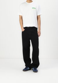 Bílá tričko s textem "Na dovolené" v zelené barvě, spárováno s černými širokými kalhotami a modrými teniskami s světle modrými pruhy.