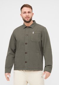Chaqueta verde oliva hecha de tejido resistente, con cuello tipo solapa, botones frontales, un bolsillo en el pecho y bolsillos laterales.