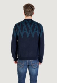 Homme avec des cheveux courts et bouclés portant un pull marine avec un motif géométrique bleu et un jean bleu, se tenant de dos sur un fond clair.