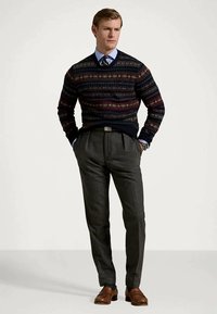 Mørkeblå mønstrede striktrøje over en lys blå skjorte, parret med grå plisserede bukser og brune lædersko. Klassisk design og teksturer.