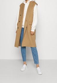 Chaleco marrón sin mangas con capucha, grandes bolsillos frontales y cierre de botones. Lleva puesto un hoodie blanco y unos vaqueros azules, combinados con zapatillas blancas.