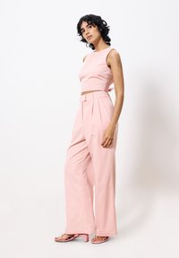 Rosa bauchfreies Top mit einer Schlaufe hinten, kombiniert mit hochgeschnittenen, weit geschnittenen rosa Hosen. Das Model trägt passende rosa Absatzsandalen.