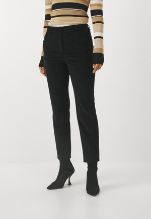 Kvinde iført sorte tætsiddende bukser, sorte ankelstøvler med hæl og en beige, sort og hvid stribet langærmet top, stående på en ensartet hvid gulv.