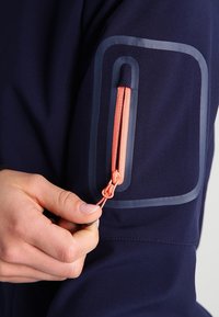 Main tirant la fermeture éclair corail sur la manche d'une veste bleue avec un galon réfléchissant foncé autour de la poche.