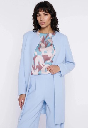 Kobieta w jasnoniebieskim płaszczu z otwartym przodem, nałożonym na wielokolorową bluzkę z abstrakcyjnym wzorem, oraz pasujące jasnoniebieskie spodnie o szerokich nogawkach.