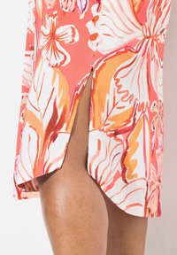 Blommig klänning med ett livfullt korall- och vitt mönster med blad- och blomdesign, kort sidospalt och mjuk tygstruktur.