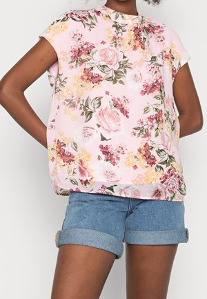 Blusa floreale rosa con maniche corte, decorata con rose, fiori gialli e foglie verdi. Abbinata a pantaloni corti in denim arrotolati.