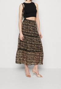 Schwarzes Cropped Top, gemusterter langer Rock mit gestuften Volants in Brauntönen, Orange und Creme. Klare High-Heel-Sandalen runden den Look ab.
