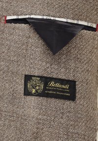 Brauner Fischgrätstoff mit einem schwarzen Etikett, auf dem "BELLANDI" und "AUTHENTIC ITALIAN FABRIC" steht. Mit einer kontrastierend abgesteppten Tasche.