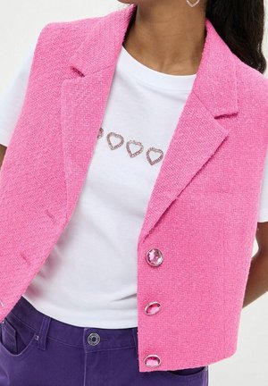 Frau trägt einen strukturierten pinkfarbenen ärmellosen Blazer über einem weißen Hemd mit herzförmigen Schmucksteinen, kombiniert mit dunkelvioletten Hosen.