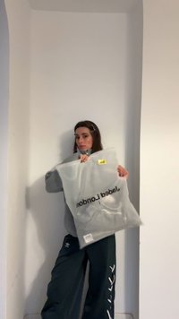 Kvinde i grå sweater og sorte Adidas-bukser holder gennemsigtig mærkevarepose med teksten "Jaded London" mod en ensfarvet hvid væg.