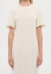 Robe droite de couleur crème avec manches courtes, encolure ronde et fente latérale. Texture lisse, fabriquée dans un tissu léger et fluide.