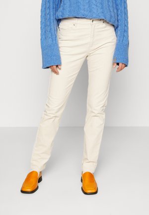 Osoba ubrana w kremowe spodnie, pomarańczowe buty z kwadratowymi noskami i niebieski sweter warkoczowy stojąca na białym tle.