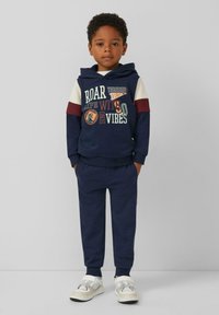 Marineblauer Hoodie mit Ärmeln in Mischfarben, der Dinosauriergrafiken und Text zeigt. Kombiniert mit passender Hose und weißen Sportschuhen.
