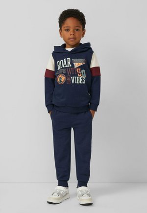 Marineblauwe hoodie met mouw in verschillende kleuren, voorzien van dinosaurustekeningen en tekst. Gecombineerd met bijpassende broeken en witte sportschoenen.
