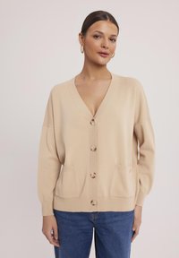 Beżowy sweter z dzianiny z dekoltem w serek, pięcioma guzikami z przodu, dwiema przednimi kieszeniami i luźnymi rękawami.