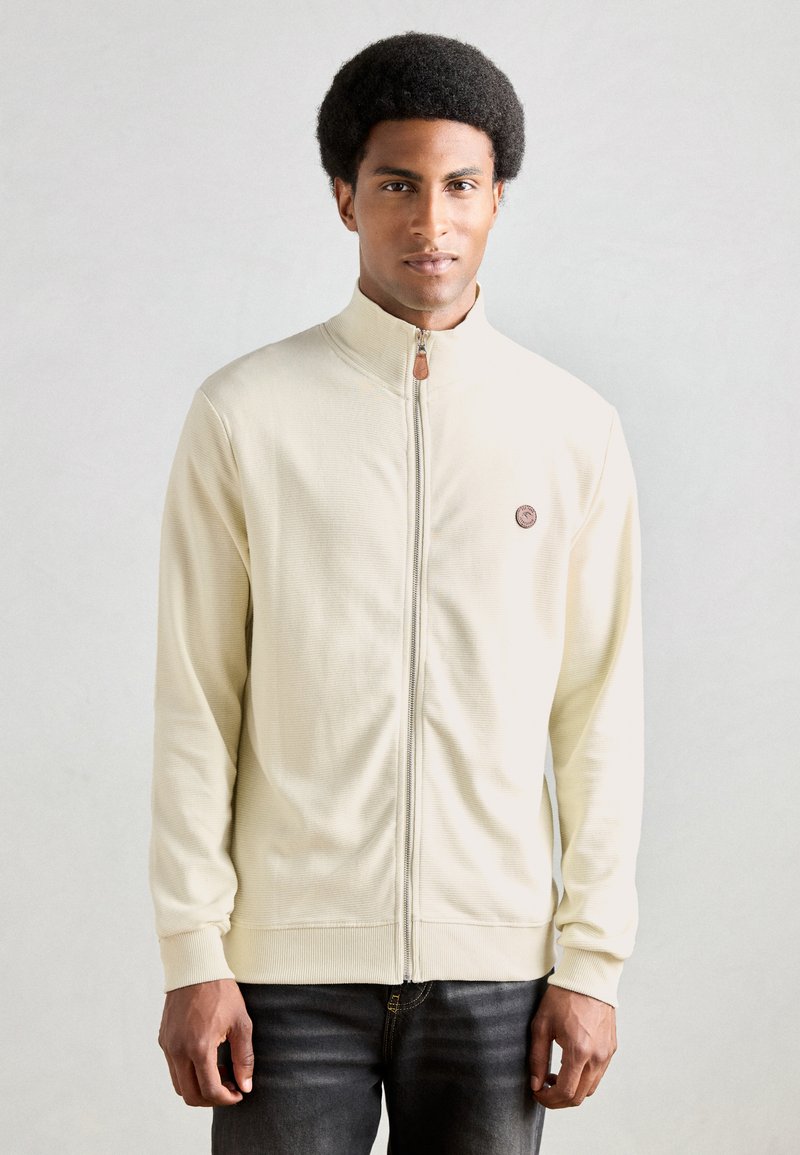 Ritssluiting lichtbeige jack met een hoge kraag en ribbelstructuur, met een klein embleem op de linkerborst en aansluitende manchetten.