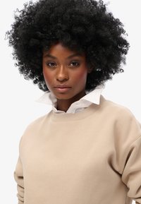 Beige sweatshirt med rund hals og ribbede manchetter, lagd over en hvid kravebluse. Har en blød tekstur og et afslappet design.