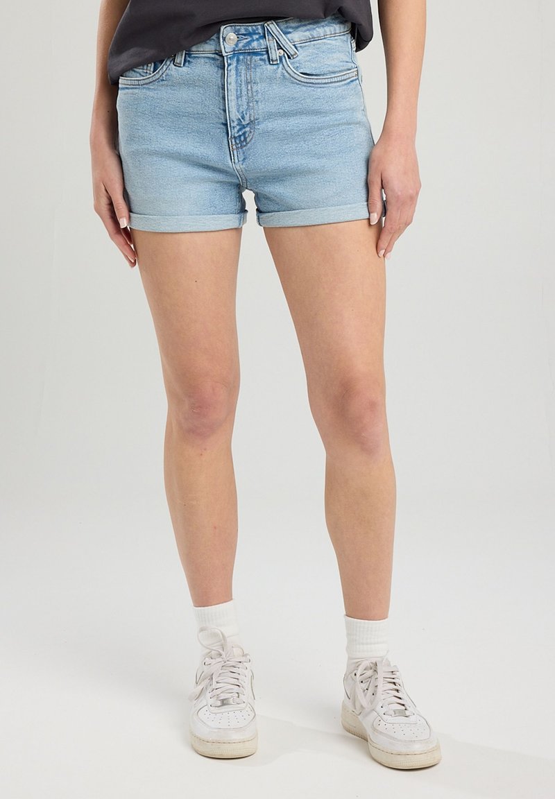 Vaaleansiniset farkkushortsit, joissa on klassinen leikkaus, käännetyt lahkeet ja viisi taskua, yhdistettynä valkoisiin lenkkareihin ja nilkkasukkien kanssa.