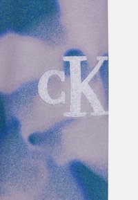 Close-up van paarse stof met een blauwgroen camouflagepatroon en witte verhoogde geborduurde letters "cK".