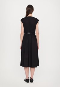 Abito nero con maniche corte a stampa, vita arricciata e gonna fluente; presenta una cintura nera lucida con borchie; indossato con scarpe casual.