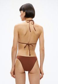 Γυναίκα με κοντά σκούρα μαλλιά που φοράει καφέ μπουστάκι �μπικίνι δετό στον λαιμό και αντίστοιχα κάτω μέρη, γυρισμένη προς τα πίσω μπροστά σε λευκό απλό φόντο.