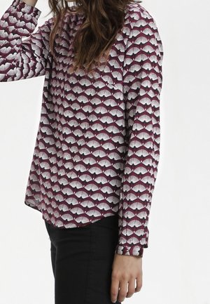Femme portant une blouse bordeaux à manches longues avec un motif blanc en forme d'éventail et un pantalon noir, vue de côté avec le bras levé vers la tête.